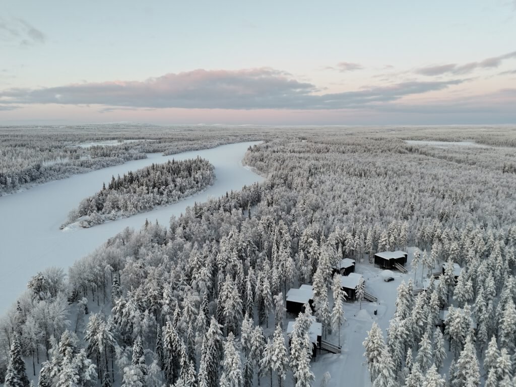 Aerial winter landscape with NOA Villas cabins beside a frozen river and snow-covered forest.