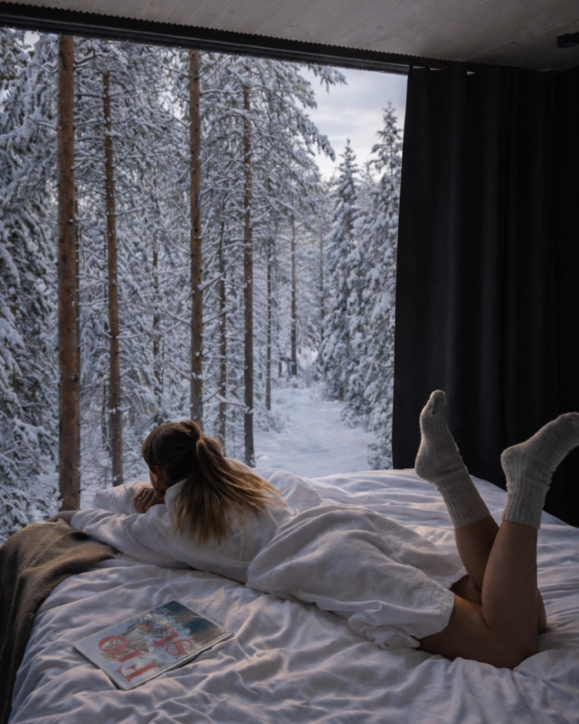 Une personne se détendant sur un lit près d’une fenêtre donnant sur une forêt enneigée.