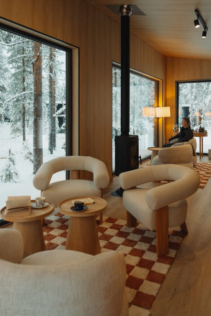 Cozy villa lounge with armchairs, tea service and snowy forest views through large windows.